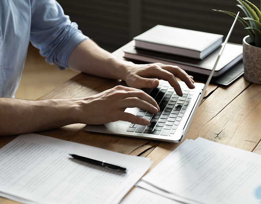 Close up businessman using laptop, typing on keyboard, sitting at wooden desk with documents